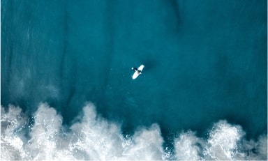 Ariel image of a surfer on white surfboard approaching a crashing wave